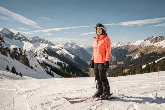 Skieuse en veste corail sur une piste printanière des Sept Laux avec panorama alpin en arrière-plan